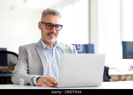 Reifer, gutaussehender, grauhaariger Geschäftsmann in einem intelligenten Blazer, der fleißig an seinem Laptop arbeitet und die Fokussierung und das Engagement widerspiegelt, die die heutigen Führungskräfte kennzeichnen Stockfoto