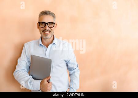 Ein entspannter, aber selbstbewusster, reifer Geschäftsmann hält seinen Laptop lässig in einem Büro mit einem frischen, modernen Design, das einen progressiven und flexiblen Arbeitsansatz anzeigt Stockfoto