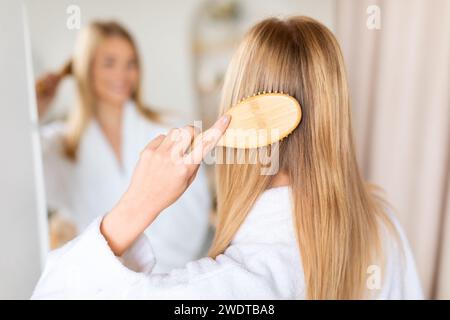 Rückansicht Frau kämmt langes blondes Haar mit einer Haarbürste im Innenbereich Stockfoto