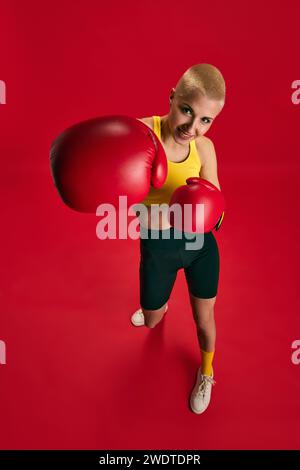 Porträt einer selbstbewussten Boxerin mit gelbem Oberteil, schwarzen Shorts und roten Handschuhen, die vor der Kamera vor rotem Hintergrund boxen. Stockfoto