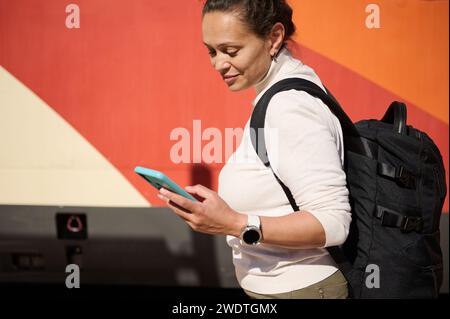 Nahaufnahme Porträt multirassische Pendlerin mit Koffer und Rucksack, Handy halten, wegschauen, auf der Reise am Bahnsteig stehen Stockfoto