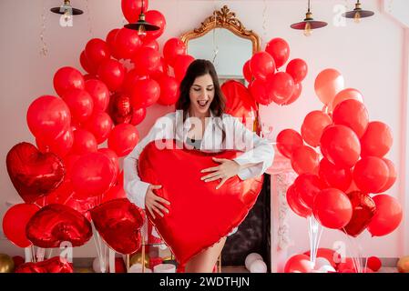 Junge Frau lächelt, während sie im Zimmer steht, das für Valentinstag dekoriert ist. Das Mädchen hält einen großen Ballon in Form eines roten Herzens. Rosenblätter verstreut Stockfoto