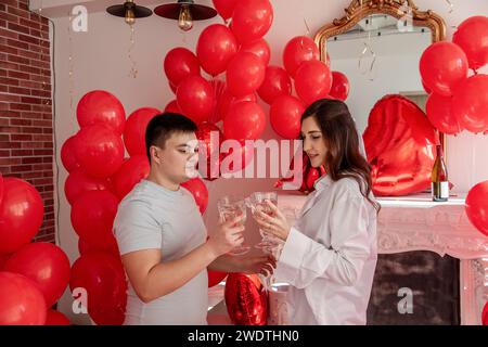 Fröhlicher Moment zwischen jungen Paaren, die mit Toast den Valentinstag in der Nähe von roten Ballons und weißem Kamin feiern. Frau lacht, hält Glas, kurz vor C Stockfoto