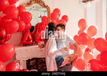Fröhlicher Moment zwischen tanzendem jungen Paar, das mit Toast den Valentinstag in der Nähe von roten Ballons feiert. Frau mit Weinglas, verdeckte Augen, Männerumarmungen, Stockfoto