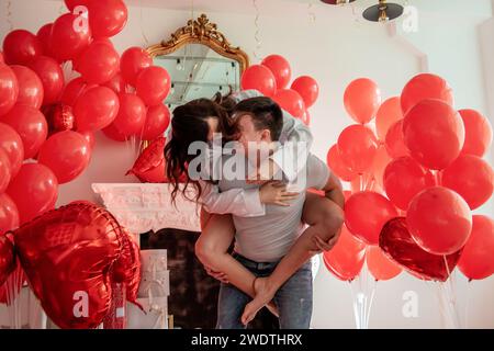 Verspielter, romantischer Moment zwischen verrücktem Paar in der Nähe von roten Ballons in einem hellen Raum mit weißer Inneneinrichtung. Frau sitzt auf dem Rücken des Mannes. Ein paar, das herumspielt Stockfoto