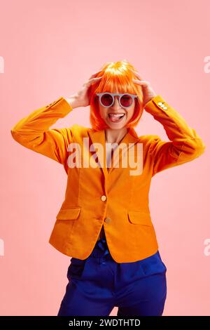 Porträt einer Frau in leuchtendem orangefarbenem Blazer, blauer Hose und orangefarbener Perücke mit Sonnenbrille, die Freude vor rosa Hintergrund ausdrückt. Stockfoto