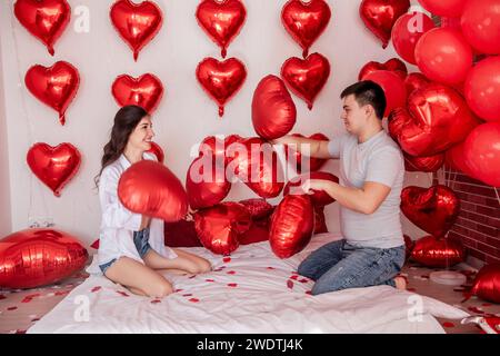 Verspielter Moment zwischen Paaren, die sich in Kissenballons auf dem Bett kämpfen, mit herzförmigen roten Ballons im Hintergrund. Die Frau macht fröhlich rumtäuscht Stockfoto