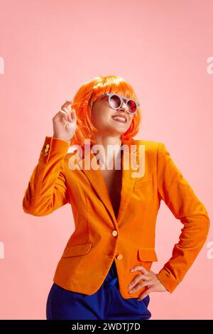 Junge träumende Frau in leuchtend orangefarbener Perücke und Blazer, in Retro-Sonnenbrille, die vor pastellrosa Hintergrund wegschaut. Stockfoto