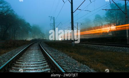 Die Eisenbahn an einem nebeligen Abend. Verschwommene Lichter eines vorbeifahrenden Zuges. Schnell vorbeifahrender Zug. Düster kalter Abend. Stockfoto