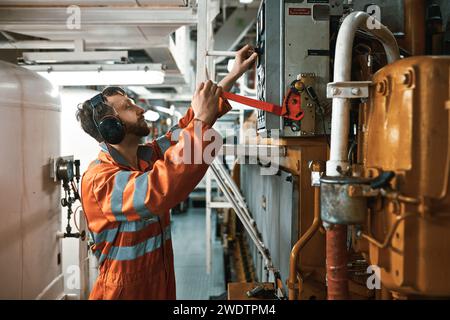 Junger Ingenieur im Maschinenraum, mit persönlicher Schutzausrüstung und manuellem Starten des Hauptdieselmotors. Stockfoto