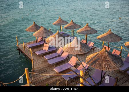 Sommerszene des Regenschirms mit Sonnenliegen auf dem Holzpier und dem Ionischen Meer in Ksamil. Ferienliege während der Goldenen Stunde in Südalbanien. Stockfoto