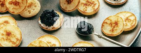 Blinis mit schwarzem Kaviar und Frischkäse Panorama, auf einem festlichen Gericht auf schwarzem Hintergrund, Mini-Pfannkuchen, eine elegante Vorspeise Stockfoto