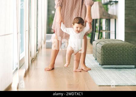 Mutter hilft dem liebenswerten Baby, ihre ersten Schritte zu machen und hält das Kind an beiden Händen Stockfoto