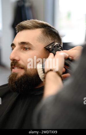 Barber rasiert die Kontur der ovalen Linie mit einem Haarschneider am Kopf des Kunden. Ein Mann mit Bart bekommt einen Haarschnitt in einem Friseurstuhl. Stockfoto