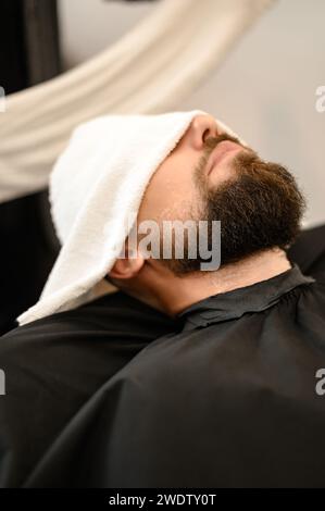 Ein Kunde liegt auf einem Stuhl in einem Friseurladen mit seinem Bart in ein Handtuch gewickelt. Ein Handtuch bedeckt die Augen des Mannes. Stockfoto