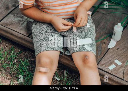 Nahaufnahme einer kindlichen Beinverletzung. Das Kind hat sich das Knie verletzt. Ein Junge legt ein Pflaster auf eine Wunde, nachdem er auf der Straße auf sein Knie gefallen ist Stockfoto