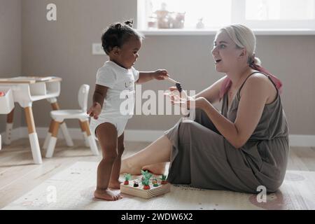 Glückliche kaukasische Mutter sitzt auf dem Boden im Kinderzimmer und spielt mit ihrer niedlichen Kleinkind-Tochter. Das Konzept der interrassischen Familie. Stockfoto