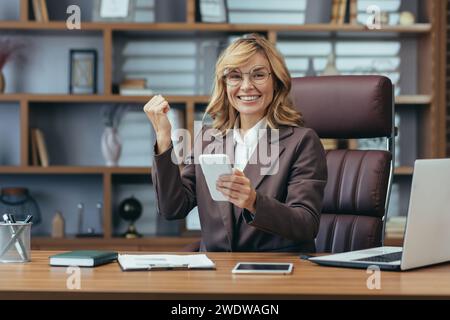 Elegante, reife, professionelle blonde Frau, die Freude ausdrückt, während sie ferngesteuert in einem stilvollen Heimbüro-Ambiente arbeitet. Stockfoto