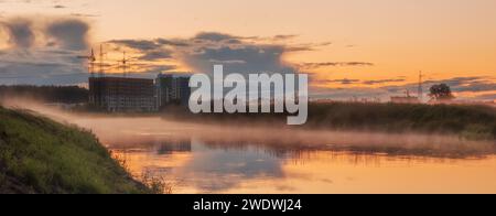 Fluss mit Nebel bei Sonnenuntergang. Wunderschöne mystische Stadtlandschaft vor dem Hintergrund eines wunderschönen bewölkten Himmels. Stockfoto