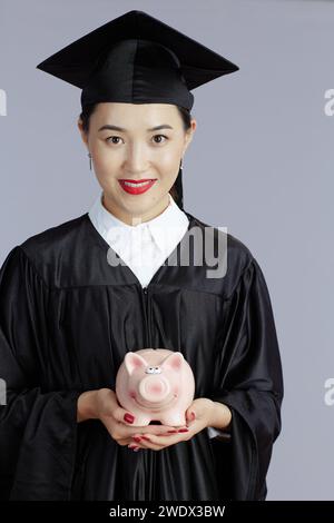 Glückliche junge asiatische Studentin mit Schweinebank isoliert auf grauem Hintergrund. Stockfoto