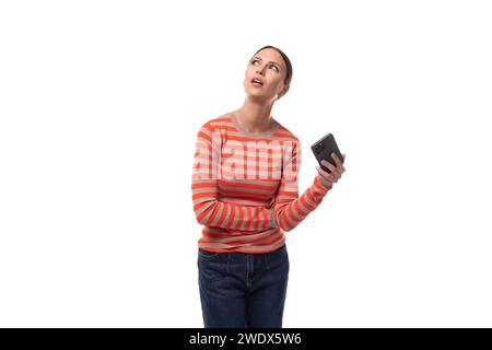 Eine junge Erwachsene Frau in einem orangefarbenen Pullover schaut träumerisch auf und hält ein Telefon Stockfoto