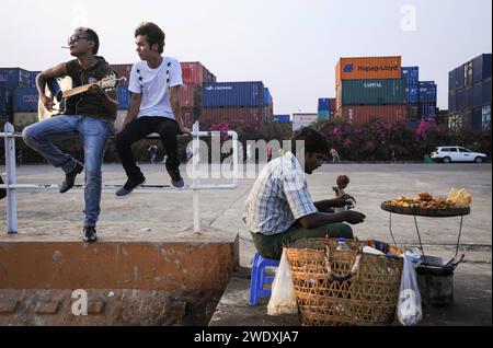 06.05.2014, Yangon, Myanmar, Asien - ein Jugendlicher spielt am Ufer des Yangon River Gitarre und raucht dabei eine Zigarette, waehrend neben ihm ein Strassenhaendler kleine Imbisse verkauft. Im Hintergrund Stapeln sich Frachtcontainer. *** 06 05 2014, Yangon, Myanmar, Asien Ein junger Mann spielt Gitarre und raucht eine Zigarette am Ufer des Yangon River, während ein Straßenverkäufer neben ihm Snacks verkauft Stockfoto