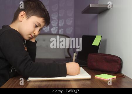 Online-Fernunterricht. Schuljunge studiert zu Hause und macht Hausaufgaben. Gelangweilter Student, der versucht, Komposition zu schreiben. Bleiben Sie zu Hause. Stockfoto