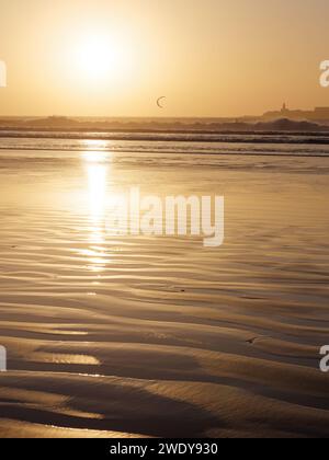 Kitesurfer auf dem Meer, während das Sonnenlicht auf dem nassen Sand bei Sonnenuntergang in Essaouira, Marokko, reflektiert, 22. Januar 2024 Stockfoto