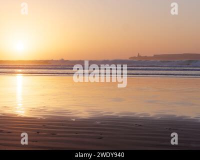 Wellen stürzen auf einen Sandstrand zu, während sich der Sonnenuntergang im flachen Wasser mit einer Insel dahinter in Essaouira, Marokko, spiegelt, 22. Januar 2024 Stockfoto