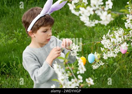 Ostereierjagd. Vorschuljunge, der Hasenohren trägt, sammelt bunte Eier auf der Ostereierjagd im Garten. Kind, das Zweige des blühenden Baumes verziert Stockfoto