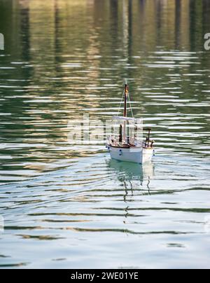 Ein Modellsegelboot begibt sich auf eine ruhige Reise über das ruhige Wasser, seine Segel entfalten sich und fangen das sanfte Licht ein. Diese bezaubernde Szene fängt ein Stockfoto