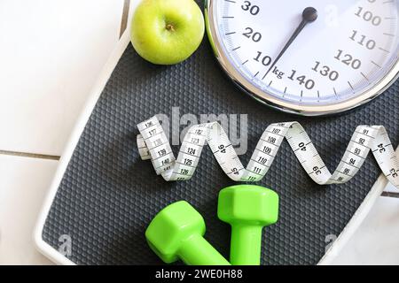 Teil einer Waage mit Maßband, grünen Kurzhanteln und einem Apfel auf einem gefliesten Badezimmerboden, Fitness- und Schlankheitskonzept, Blick von oben, Stockfoto