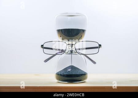 Behalten Sie die Zeit im Auge, Sanduhr mit schwarzem Sand, Brille, Geschäftskonzept der Deadline läuft ab, Holztisch vor hellgrauem Hintergrund Stockfoto
