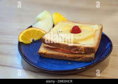 Toast Hawaii, gegrilltes offenes Sandwich mit Schinken, Ananas und Käse, ein paar Obststückchen und Himbeergarnitur auf einem blauen Teller auf einem Holztisch, Sel Stockfoto