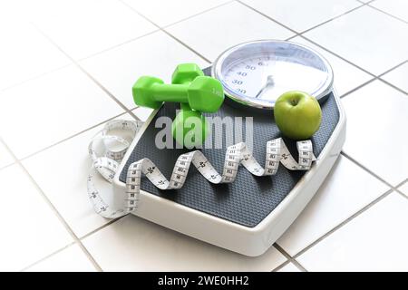 Fitness- und Gewichtsverlust-Konzept, Personenwaage mit Maßband, grüne Kurzhanteln und ein Apfel auf weiß gefliestem Badezimmerboden, Kopierraum, Auswahl Stockfoto