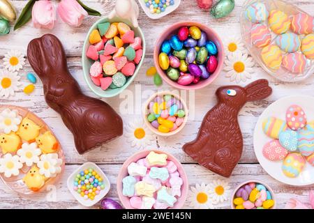 Oster-Süßigkeiten-Tischszene. Blick von oben über einen weißen Holzhintergrund. Schokoladenhasen, Süßigkeiten und eine Auswahl an Süßigkeiten. Stockfoto