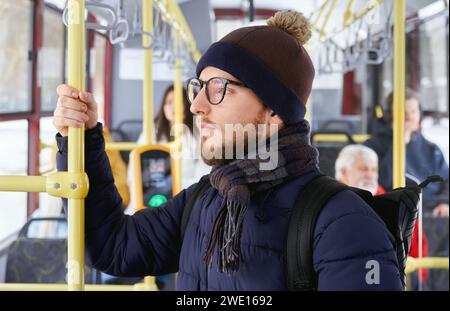 Nahaufnahme eines nachdenklichen erwachsenen bärtigen männlichen Mannes in Brille mit Rucksack, der beim Transport auf das Fenster blickt. Eine Menge junger Männer und verschwommene Passagiere, die im fahrenden Bus sitzen. Konzept des Stadtlebens. Stockfoto