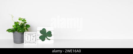 St. Patricks Day Dekor auf weißem Regal. Shamrock Pflanze, schäbiger schicker Holzkalender und Schild vor einem weißen Wandbanner. Kopierbereich. Stockfoto