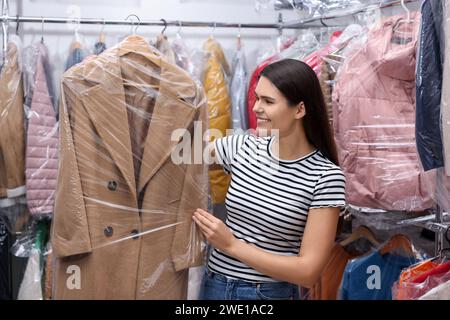 Reinigungsservice. Glückliche Frau, die einen Kleiderbügel mit Mantel in einer Plastiktüte in der Nähe von Regalen mit Kleidung im Haus hält Stockfoto