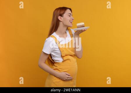 Schwangere junge Frau, die ein Stück leckeren Kuchen auf orangefarbenem Hintergrund isst Stockfoto