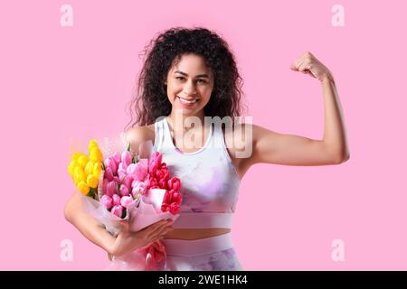 Junge afroamerikanische Frau in Sportbekleidung mit verschiedenen Blumensträußen, die Muskeln auf rosa Hintergrund zeigen. Internationaler Frauentag Stockfoto