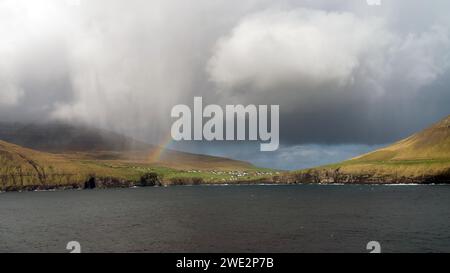 Auf den Färöern liegt ein kleines Dorf inmitten einer dramatischen Landschaft unter dem Regenbogen. Stolz erheben sich Klippen, die von der North ATL erschlagen werden Stockfoto