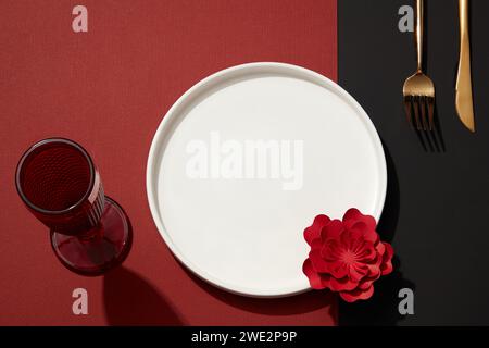 Szene mit Esstisch mit Essensutensilien. Ein runder weißer Teller, Besteck, Glasbecher und rotes Blumenpapier auf farbigem Hintergrund. Dekorat Stockfoto