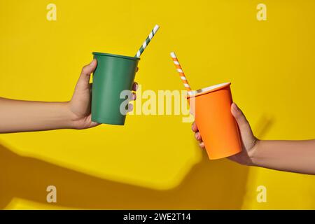 Zwei Hände von zwei Leuten, die bunte Einweg-Pappbecher mit Strohhalmen auf gelbem Hintergrund halten. Abstrakte Szene mit minimalistischem Stil für Produkt br Stockfoto