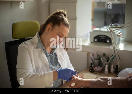 Weibliche Nageltechnikerin trägt Handschuhe, einen weißen Mantel und hält während der Arbeit die Hand ihres Kunden Stockfoto