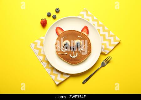 Kreatives Servieren für Kinder. Teller mit niedlicher Katze aus Pfannkuchen, Beeren, Creme, Banane und Schokoladenpaste auf gelbem Hintergrund, flach gelegt Stockfoto