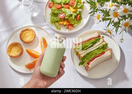 Gesunder Sommersalat vegetarisches Mahlzeitkonzept, frischer Salat mit Tomaten, Sandwiches, Orange und Kuchen auf weißem Hintergrund. Weibliche Hand hält grün B Stockfoto