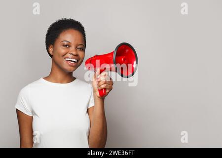 Fröhliche Frau in weißem leerem T-Shirt, das Lautsprecher vor grauem Studio-Wandbanner-Hintergrund hält Stockfoto