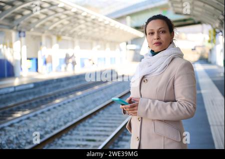Nachdenkliche junge Erwachsene Frau im beigefarbenen Wollmantel, nachdenklich in die Ferne blickt, auf einen elektrischen Hochgeschwindigkeitszug wartet, Handy in h hält Stockfoto