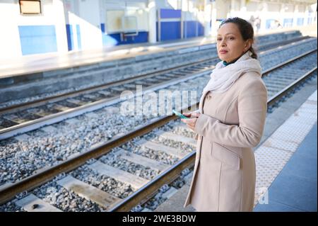 Nachdenkliche Frau, die ihr Handy überprüft, in die Ferne schaut, auf dem Bahnhof steht. Das Konzept der Menschen, des Lebensstils und der Nutzung von Hochgeschwindigkeitsgeschwindigkeit Stockfoto
